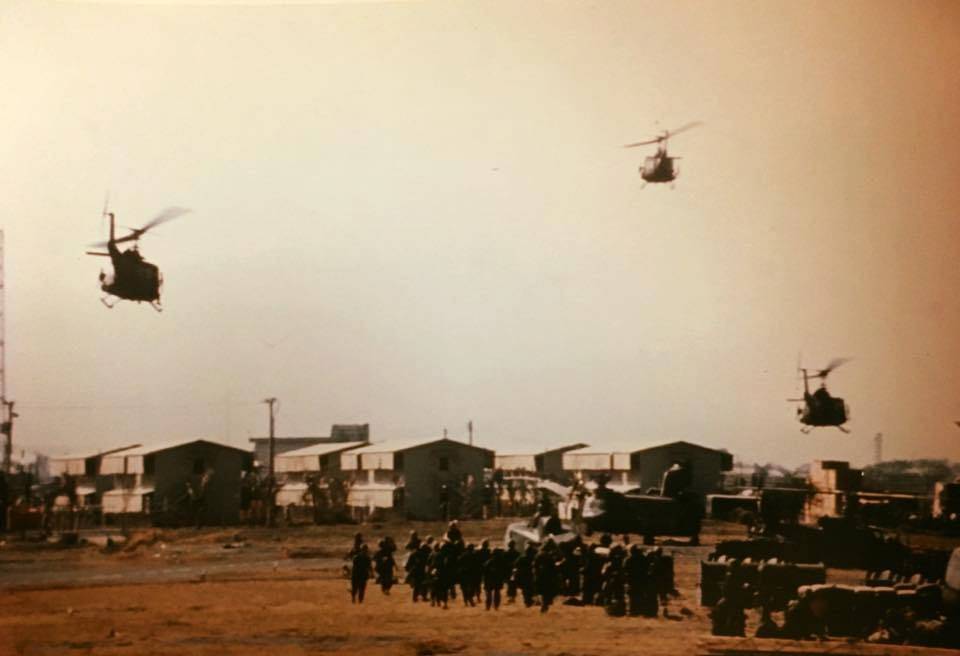 Three helicopters in the sky above a basecamp, soldiers on foot below.