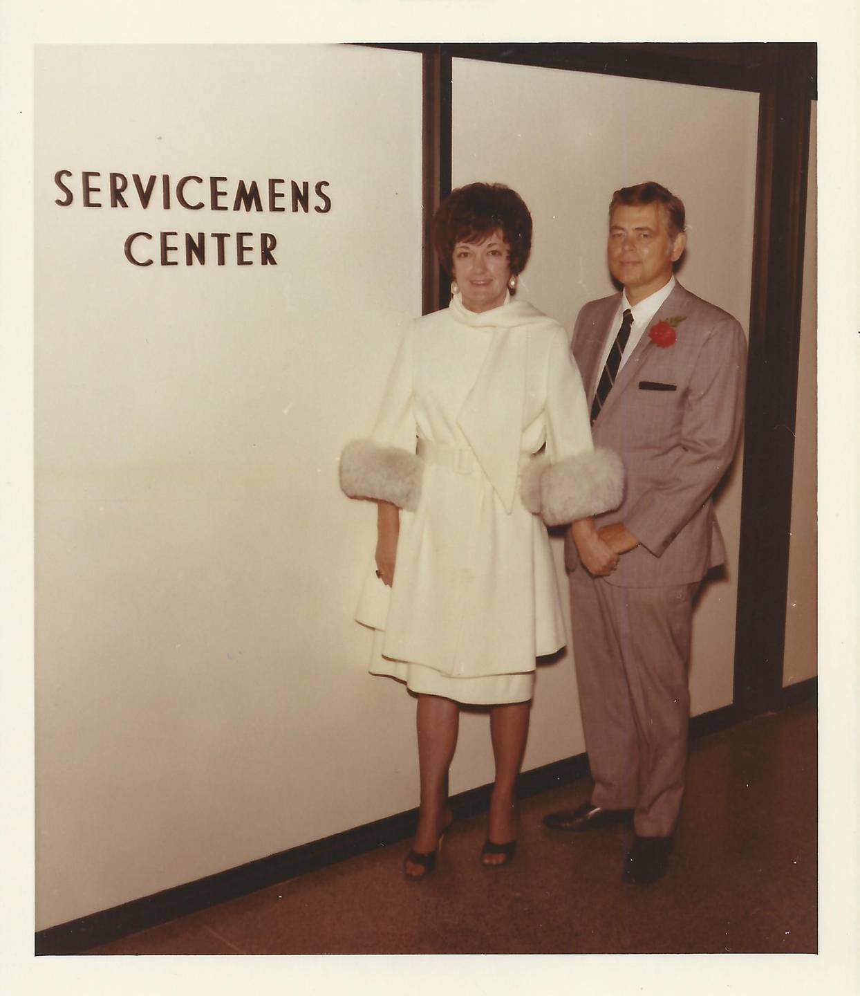 A middle aged couple dressed up, standing near a wall that reads "Servicemens Center."