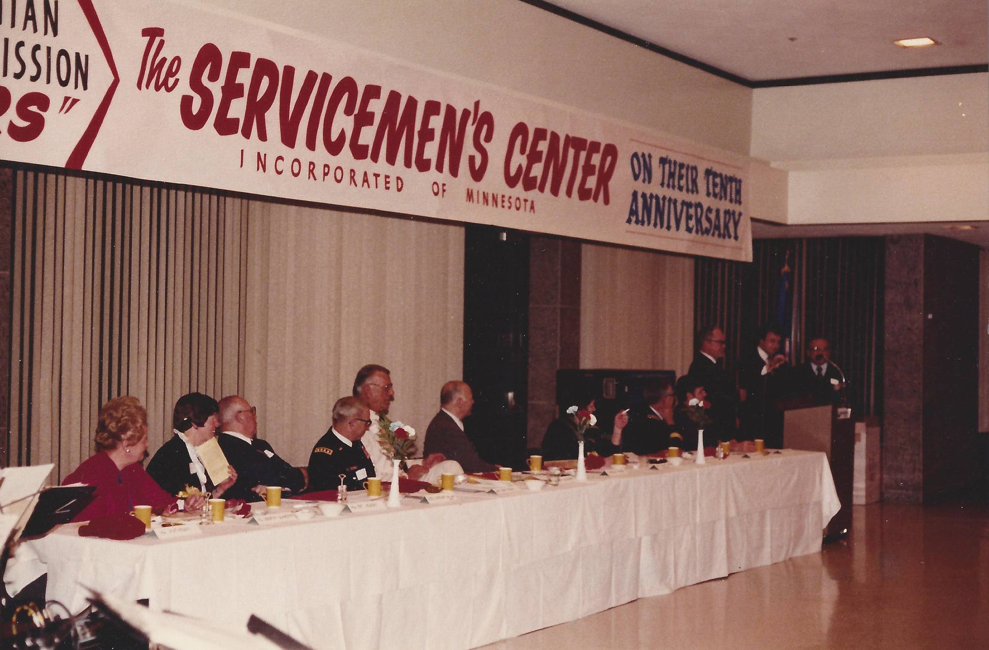 People seated at a long table with a white table cloth, looking towards a podium with a man speaking.