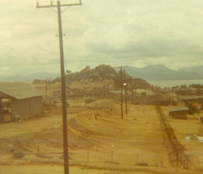 A dusty village with telephone poles rising from the ground; mountains and a bay in the background.