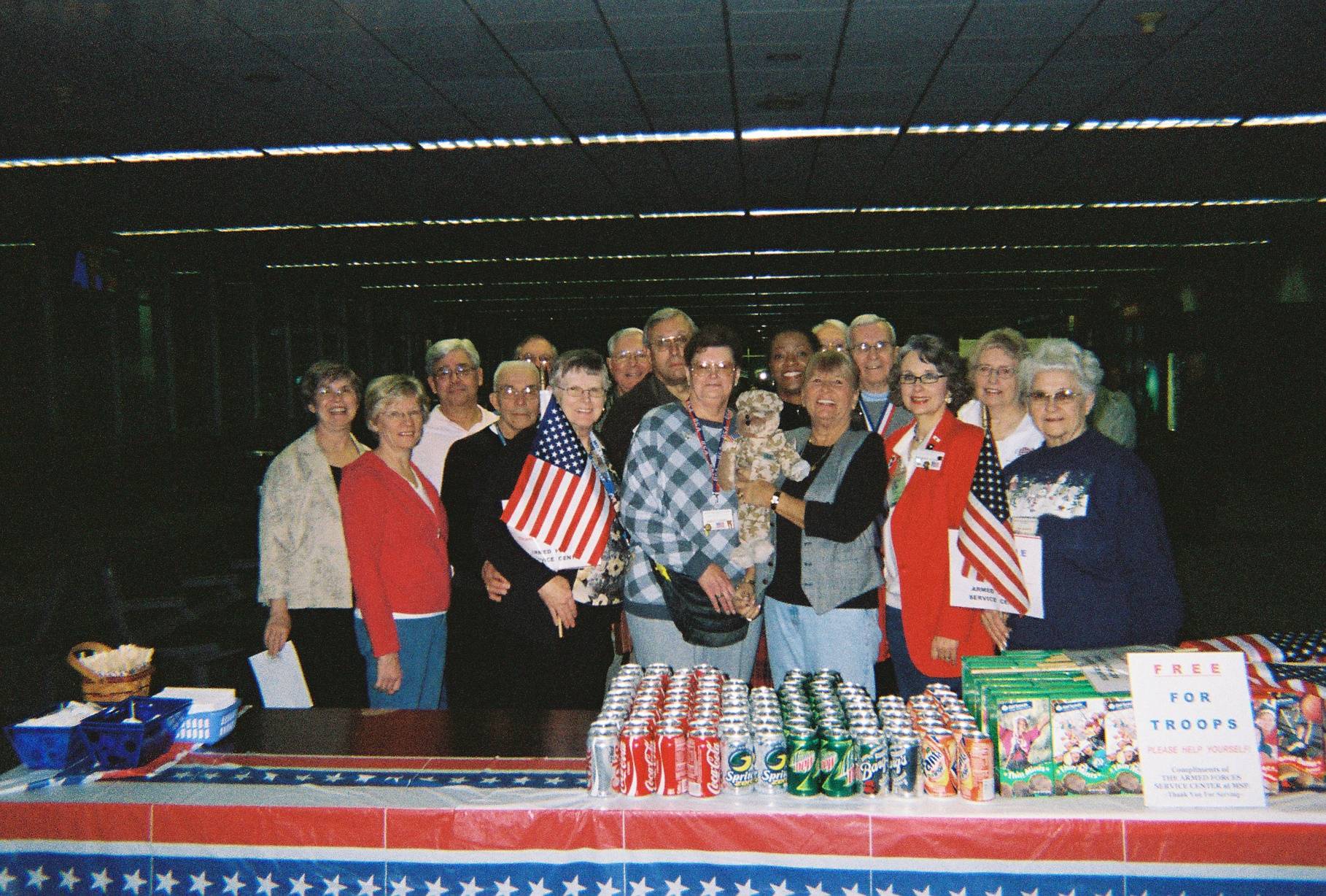 Contemporary photo of a group of people with miniature American flags, standing behind a table of refreshments.