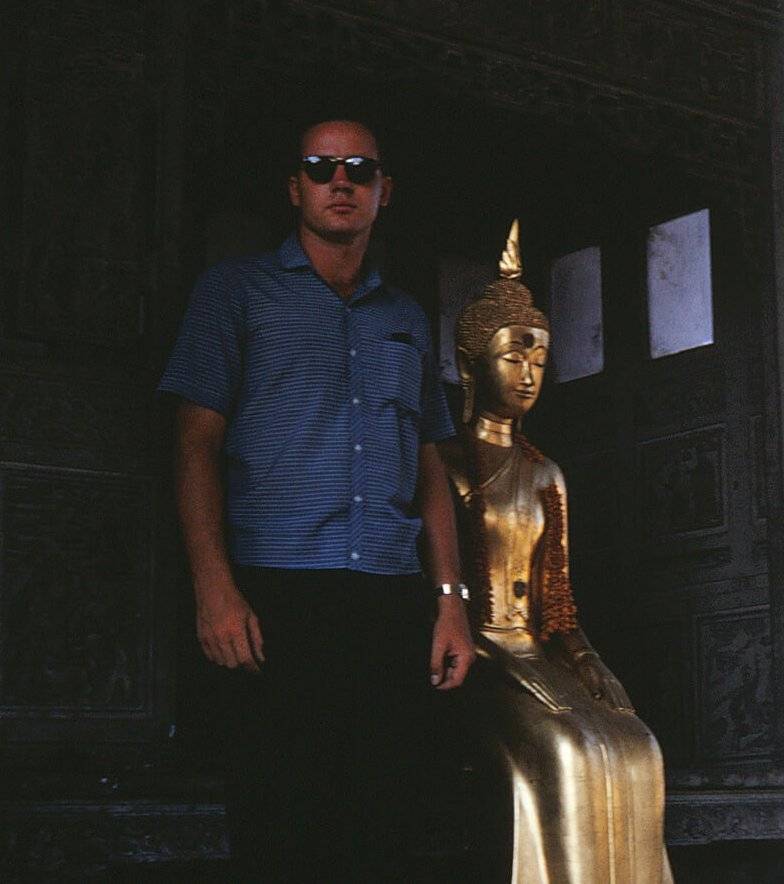 Young man in civilian clothes standing inside a temple next to a gilded statue.
