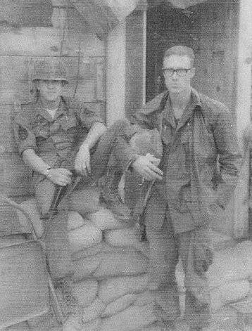 Two young men in uniform, on sitting on sandbags leaning against a building, one standing next to him with his helmet held in his arms.