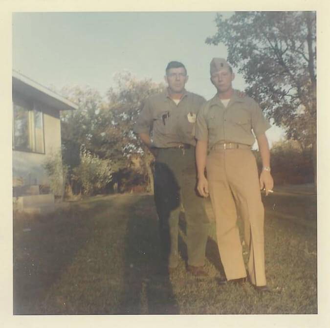 Two men -- one young, one old -- in uniform, standing in a yard outside a house.