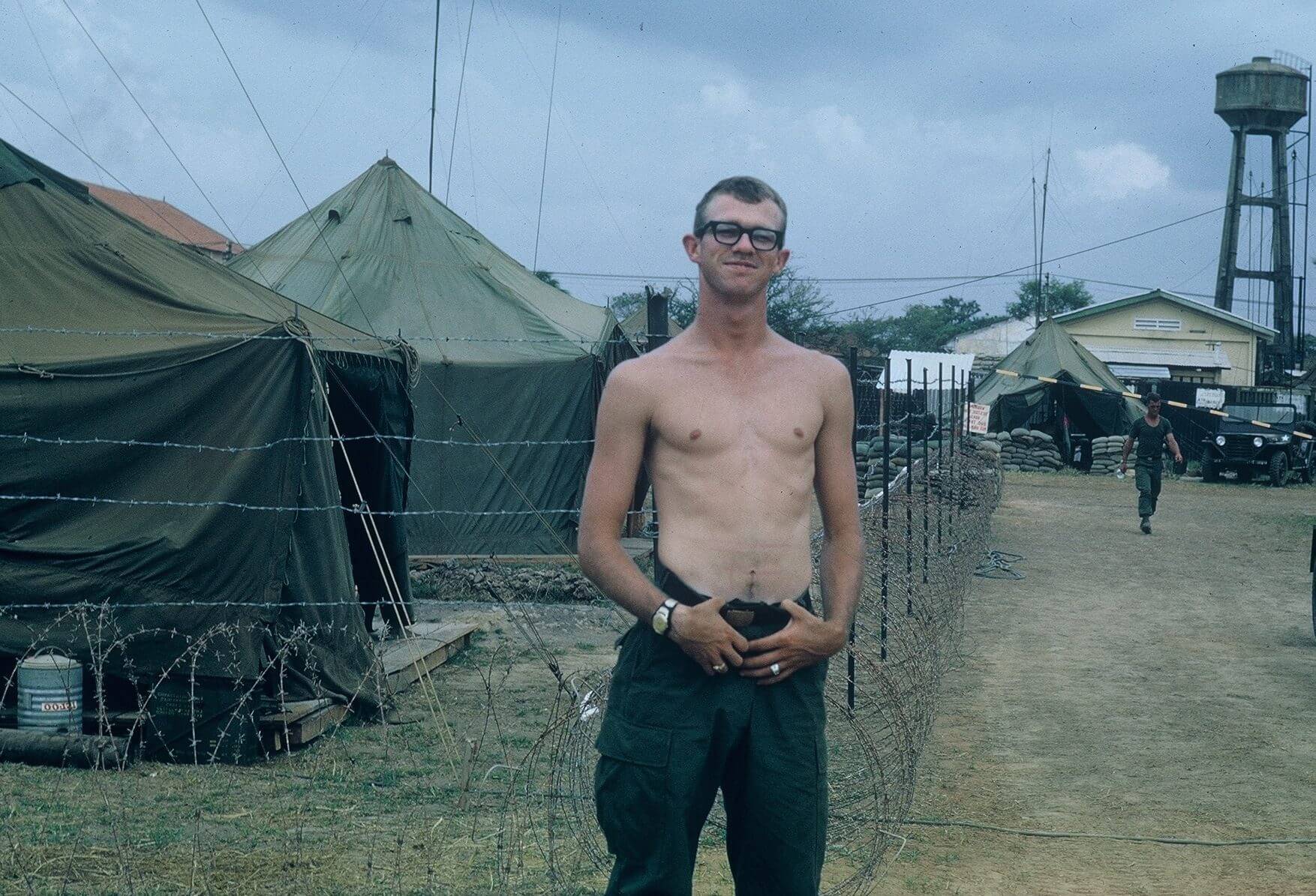 Young shirtless soldier posing outside an encampment.