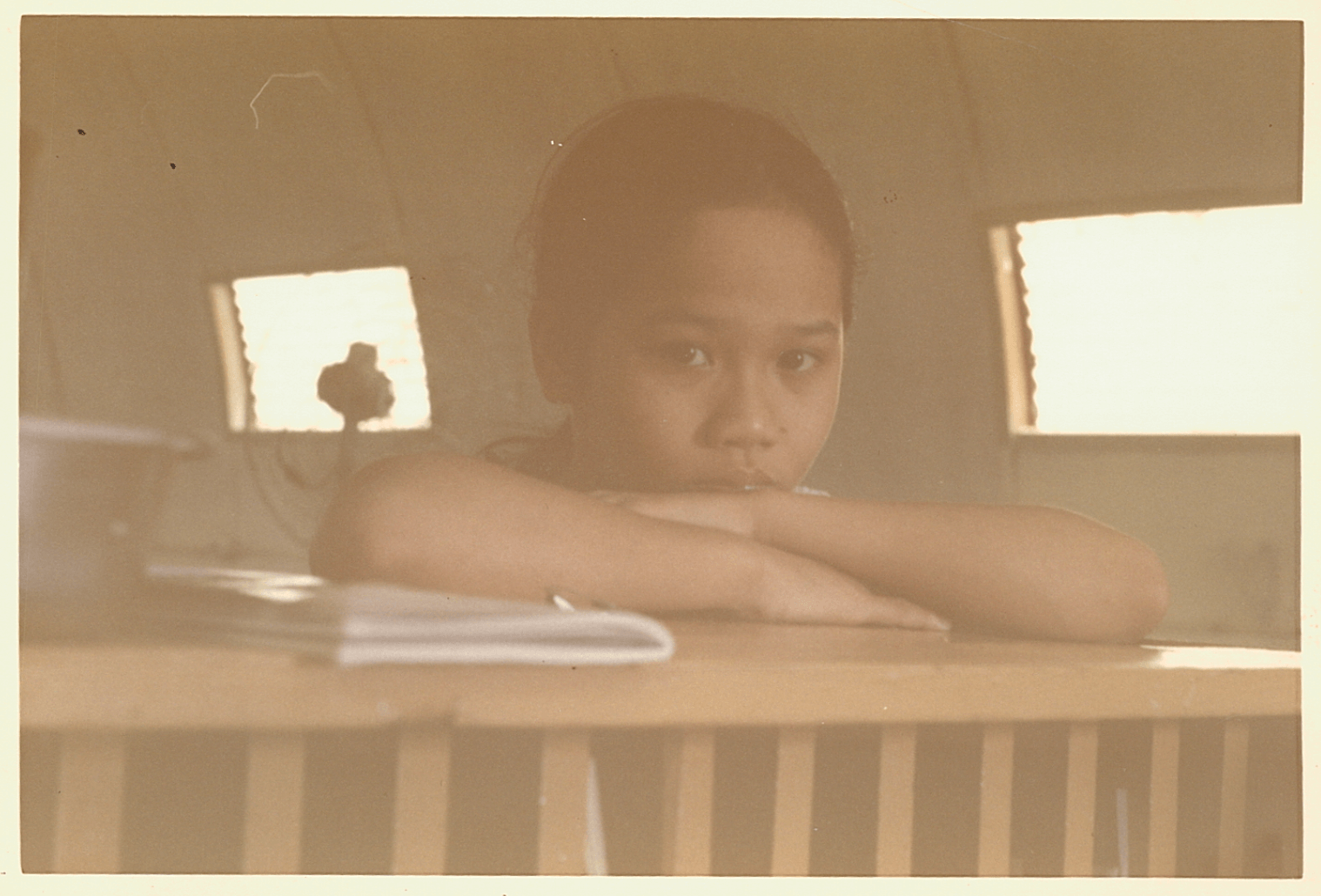 A young Asian girl peers somberly over her bed toward the camera.