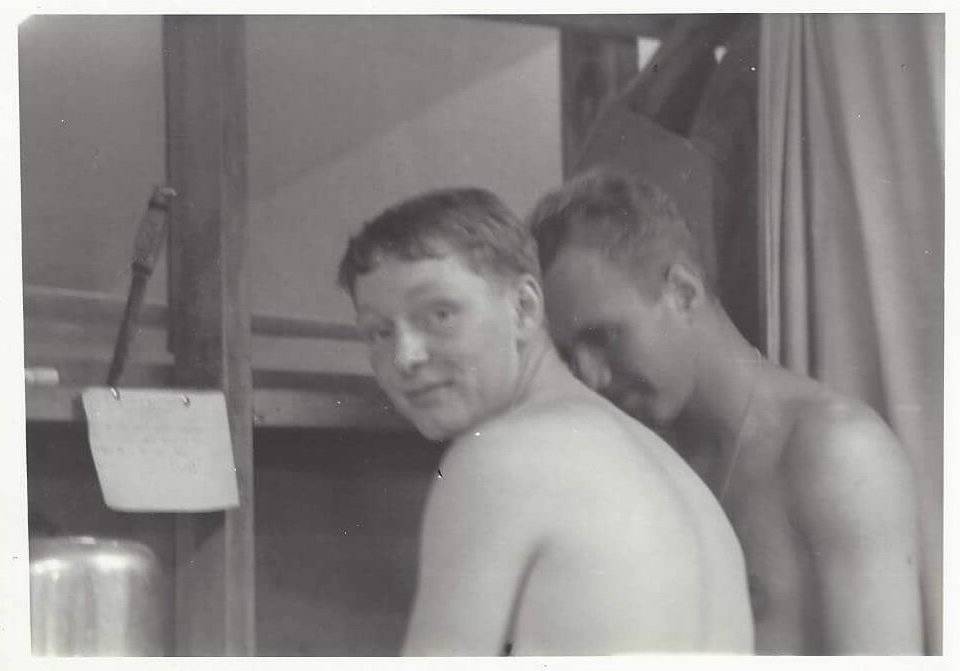 Two young, shirtless men in a barracks, the frontmost looking over his shoulder at the camera.