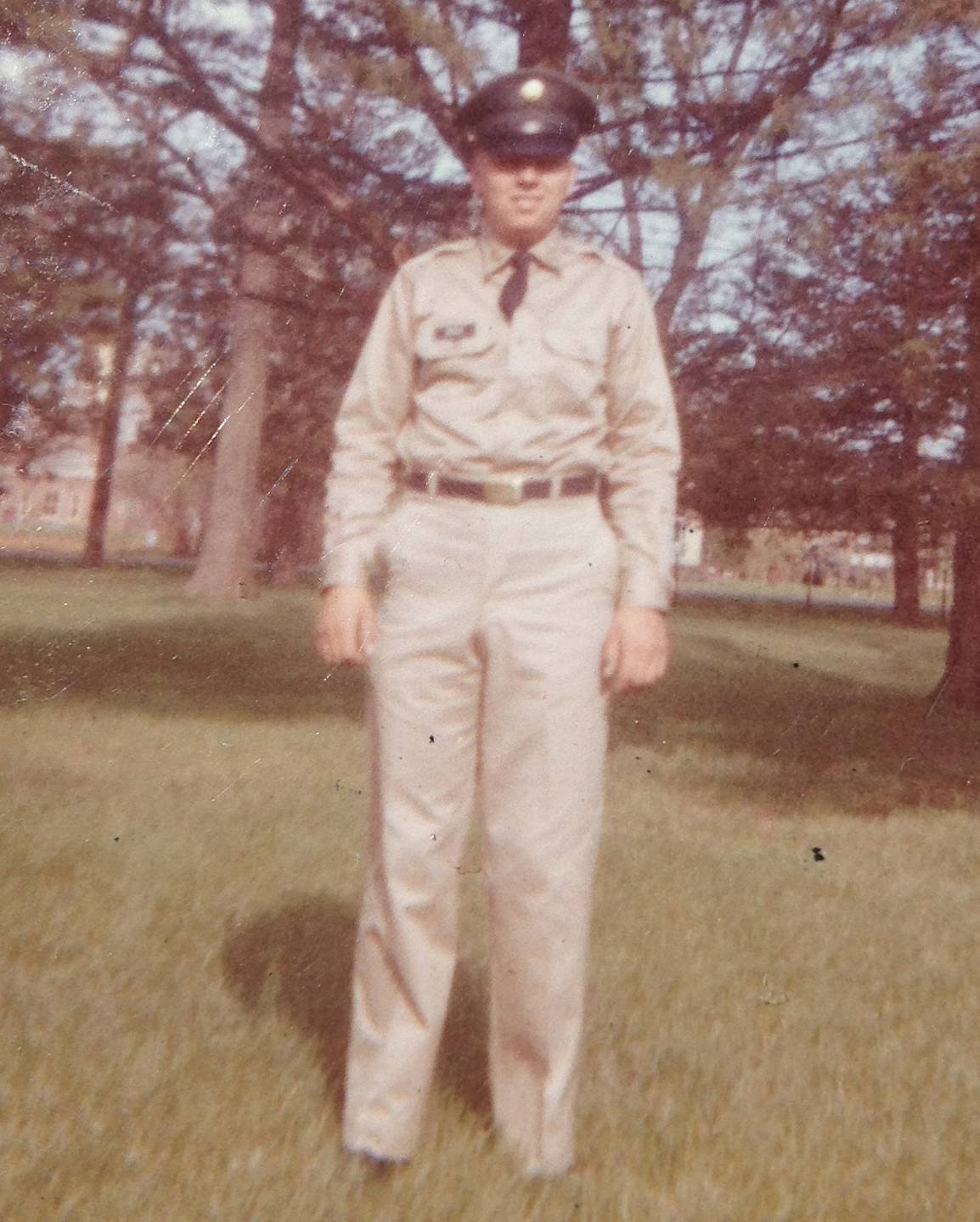 Young U.S. soldier in uniform, with hat, standing in a field of grass and trees.