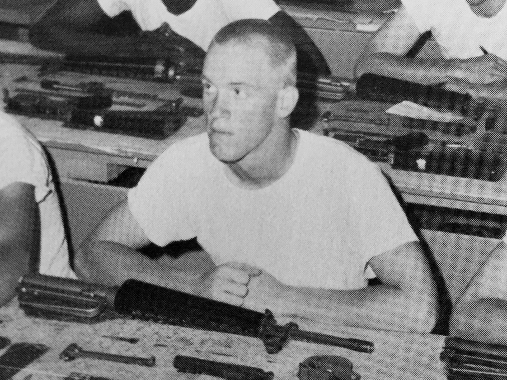 A young man in a white t-shirt at a desk with gun parts laid out in front of him.