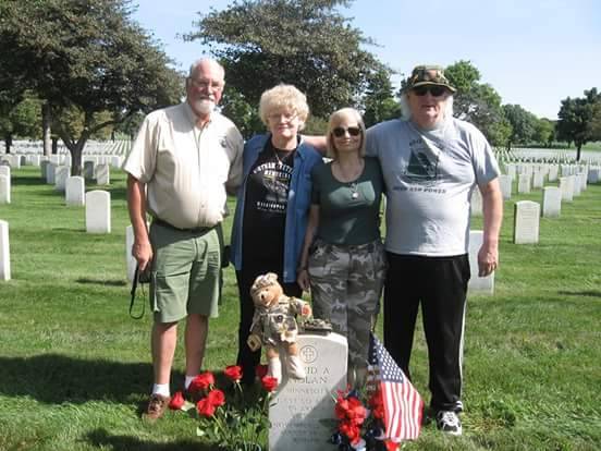 Contemporary photo of four people standing behind a white marble tombstone decorated with a miniature American flag, teddy bear in uniform, red carnations.