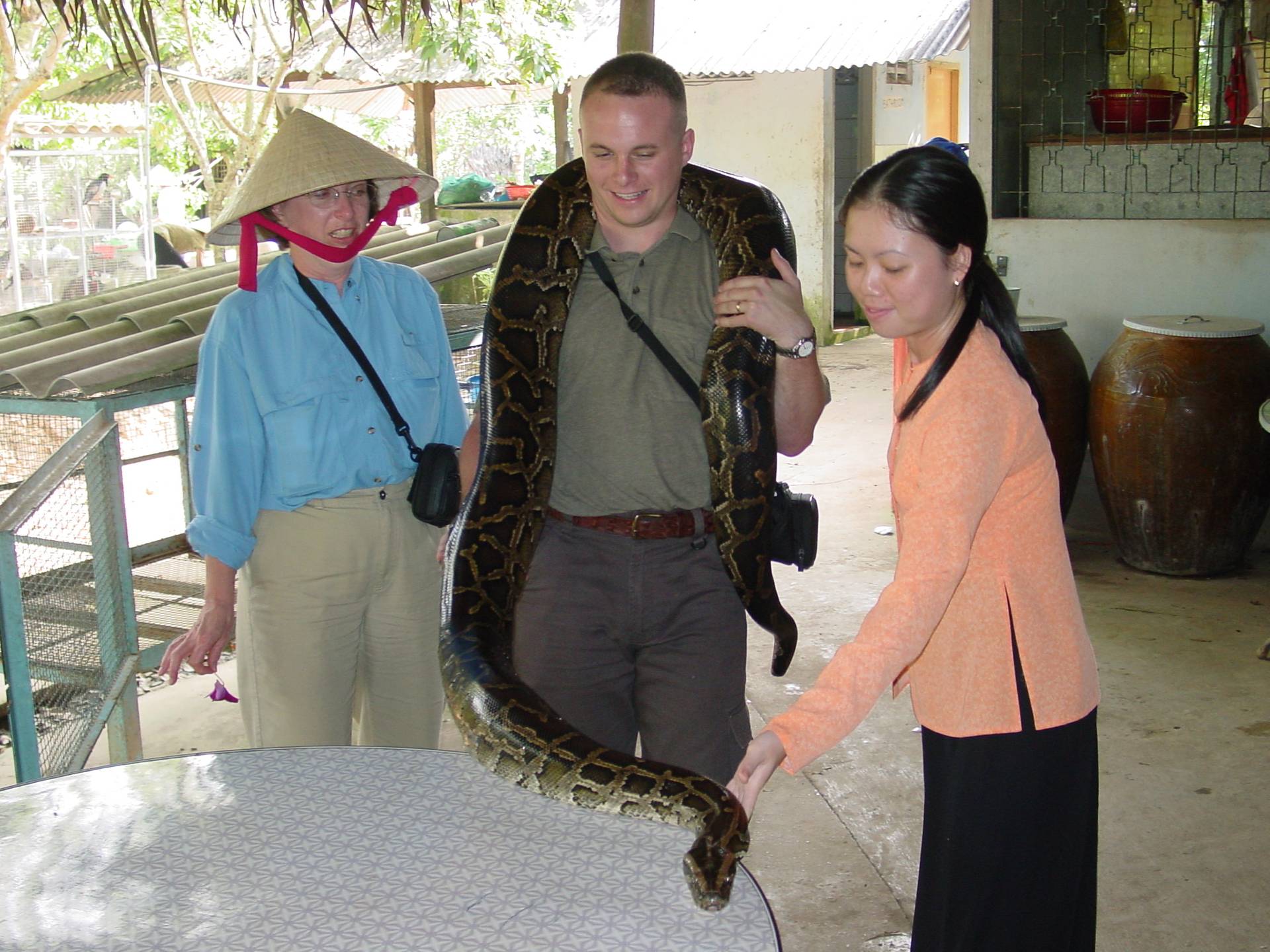 Young man with a large snake around his shoulders. Older American woman in Vietnamese straw hat and young female snake handler stand next to him.