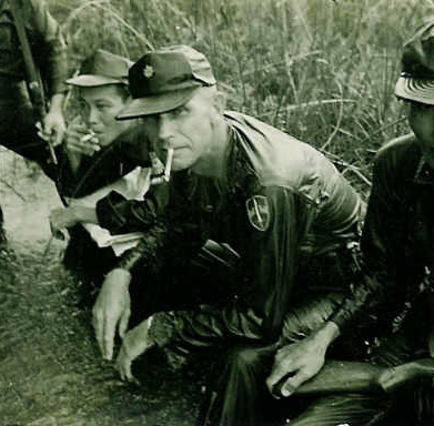 An older U.S. soldier crouched down with Asian soldiers, smoking a cigarette.