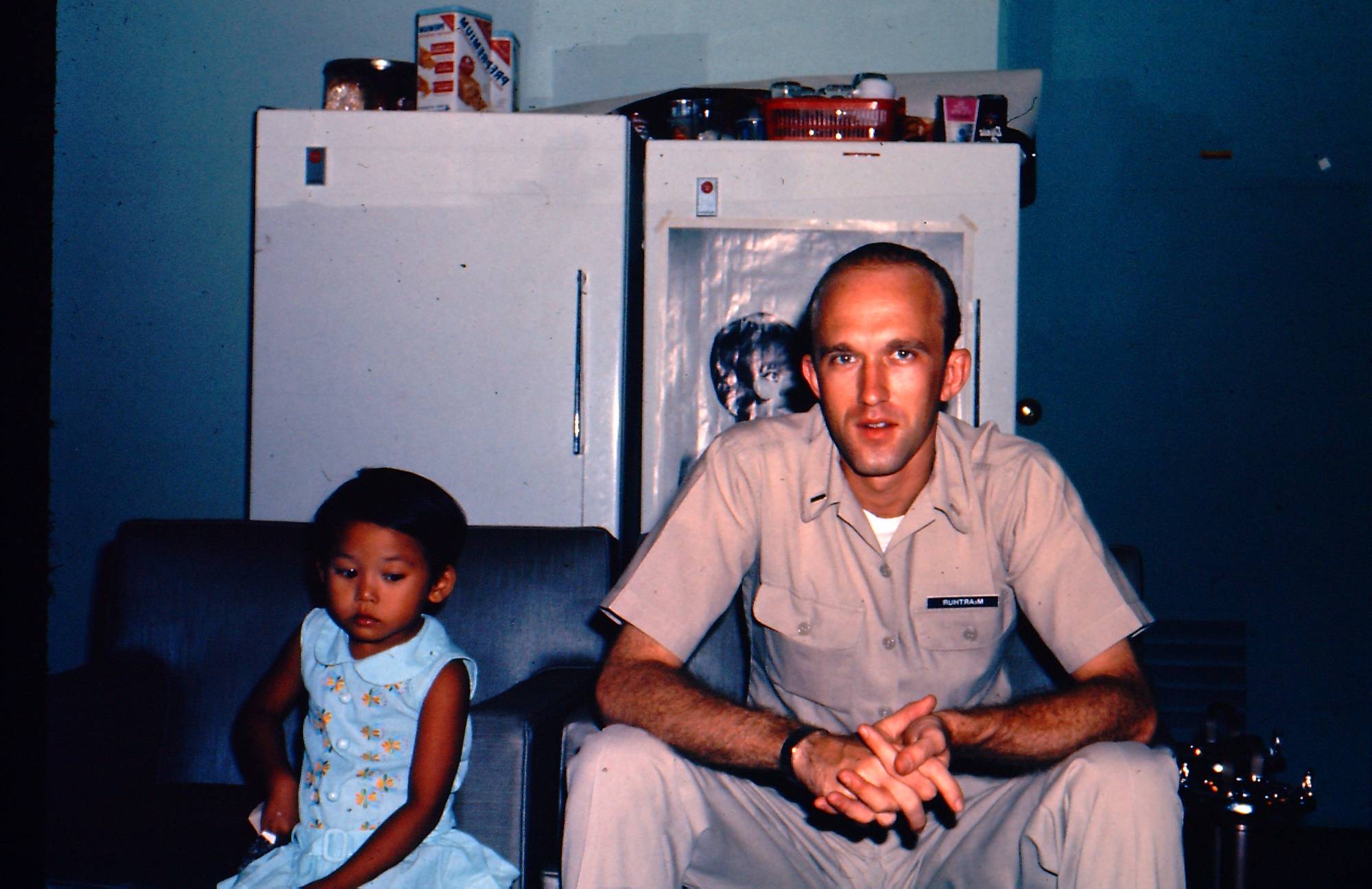 A young man in a khaki uniform seated next to a young Asian child.