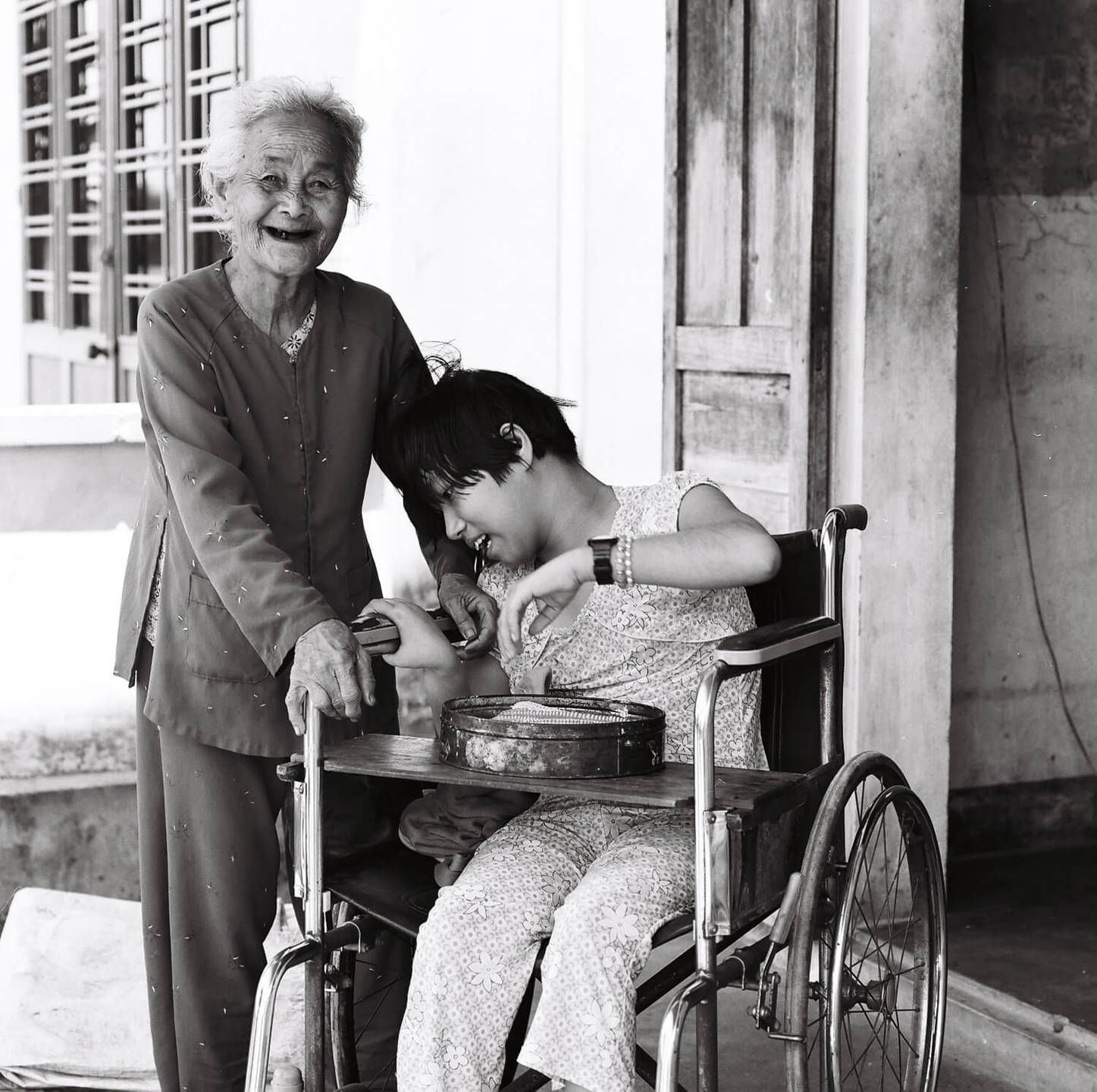 An old, smiling Asian woman standing at the side of a wheelchair-bound child.