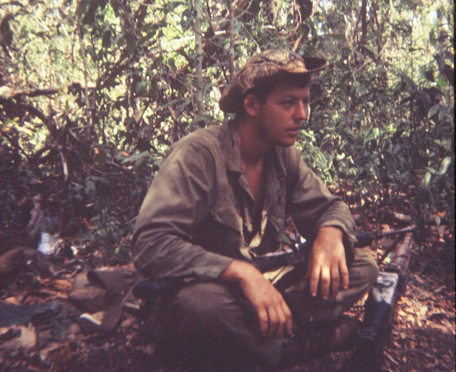 U.S. soldier in a boonie hat, sitting in the jungle with his rifle across his lap.