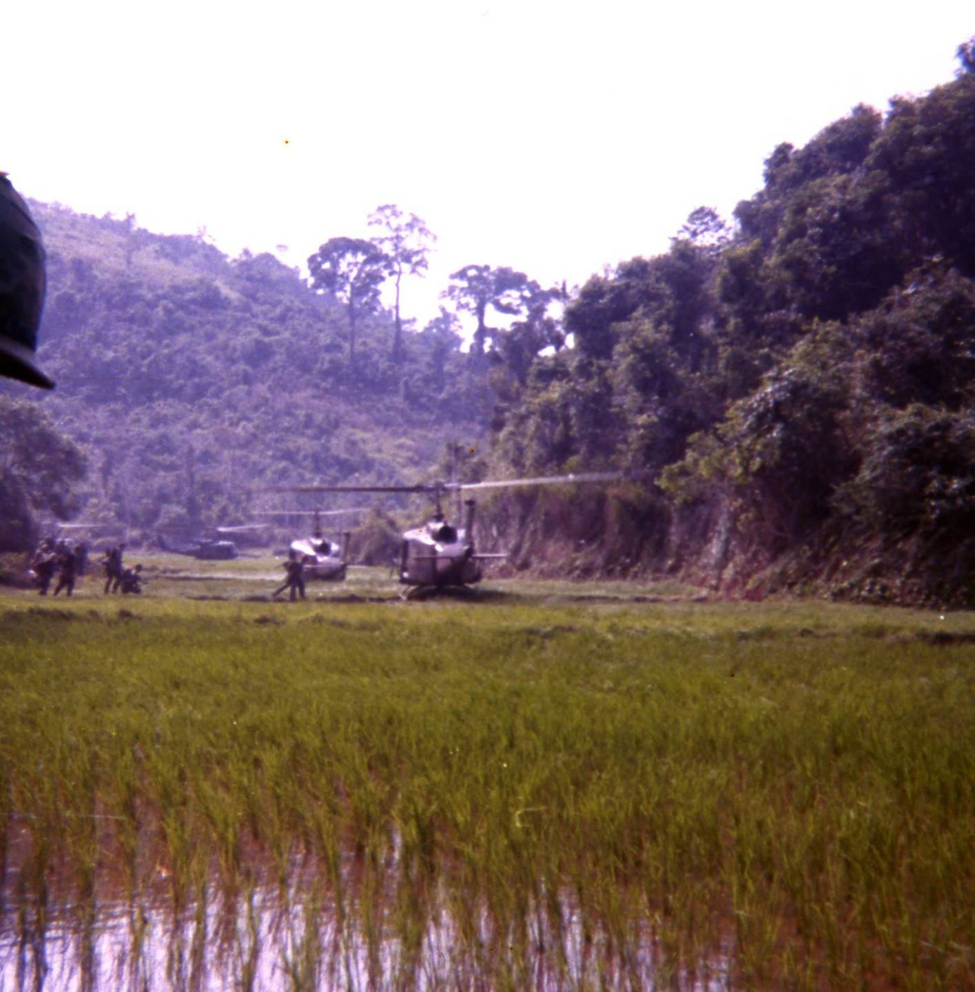 A rice paddy in a valley, helicopters on solid ground in the distance.
