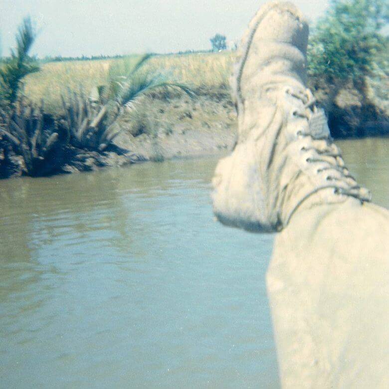 A muddy boot and pant leg, rice paddies in the background.