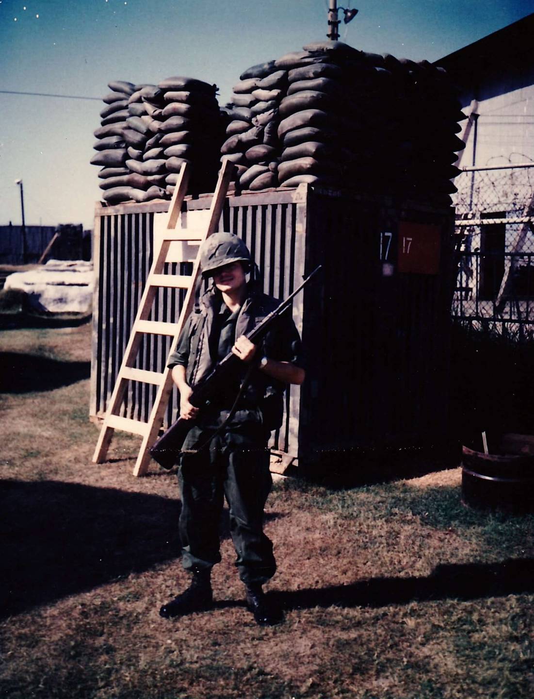 U.S. soldier in full gear, holding rifle, standing in front of a wooden structure with sandbags stacked on top of it.