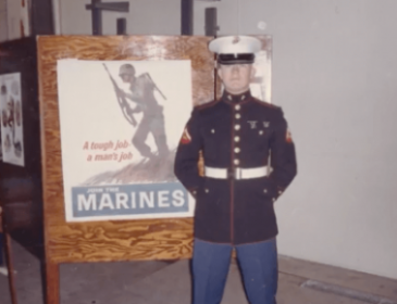 Young Marine in uniform posing in front of a Marines poster that says "A tough job, a mans job."