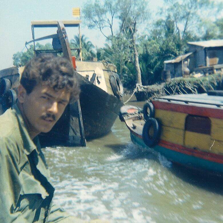 A U.S. soldier standing in the foreground, river and boats in the background.