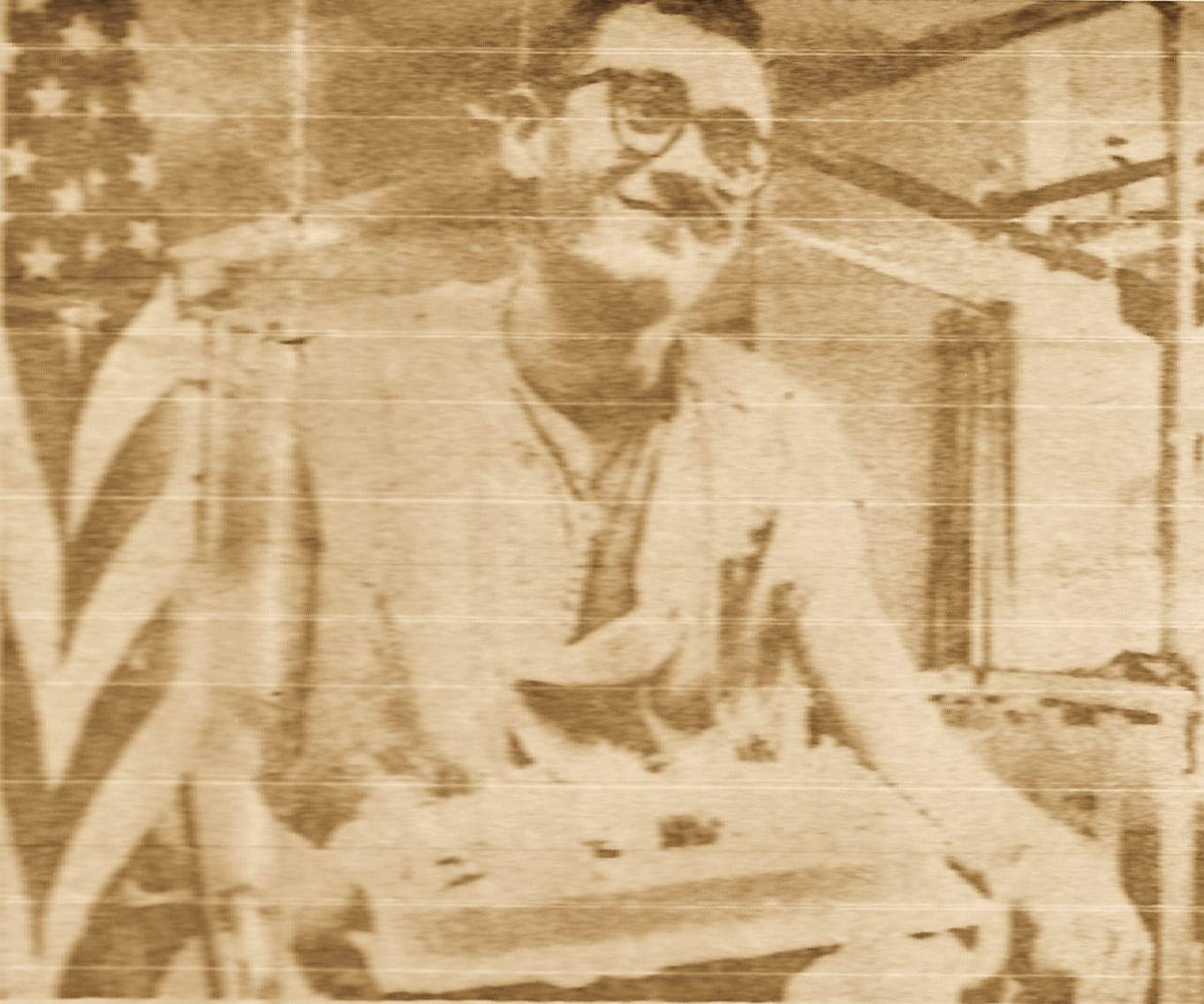 A grainy photo of a young man smiling, American flag in the foreground to the man's right.