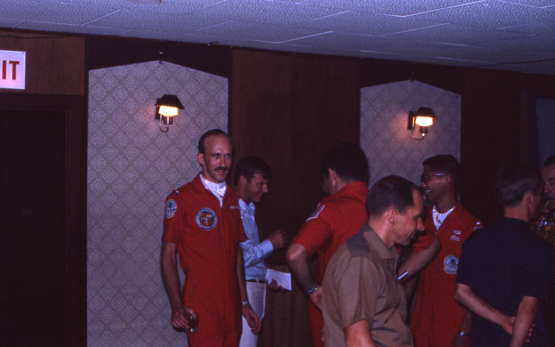 A group of men, many of them in orange flight jumpsuits, socializing in what looks to be a hotel.