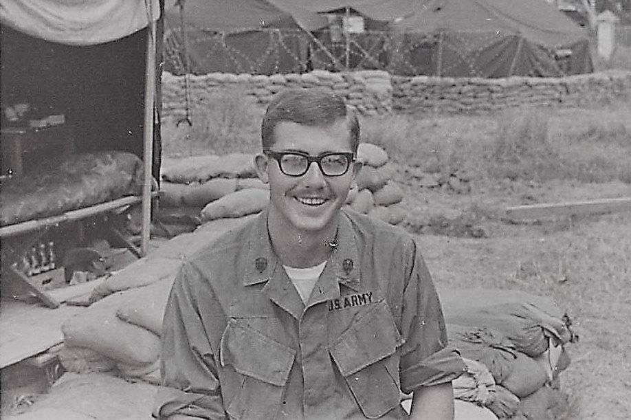 A young U.S. soldier with glasses and a mustache sitting outside an encampment on a pile of sandbags.