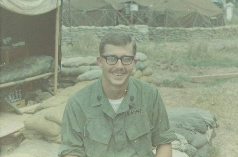 Young Caucasian soldier with mustache and glasses sitting outside among sandbags, smiling for the camera.