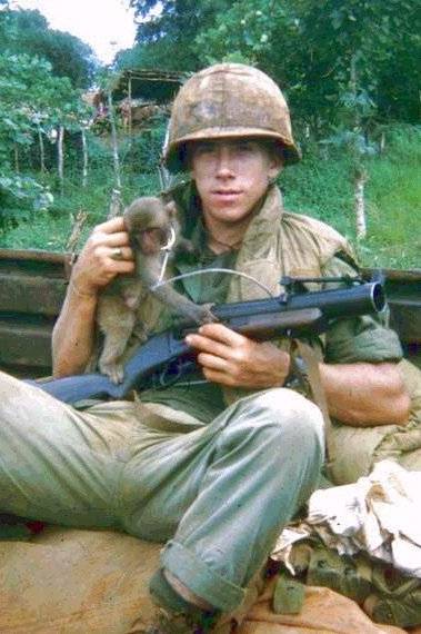 A young U.S. soldier reclined, holding a gun and a small monkey.