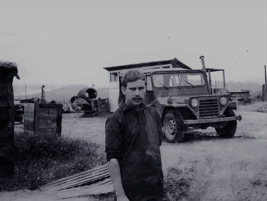 Stern-looking young U.S. soldier on a base, Jeep and small outbuilding in the background.
