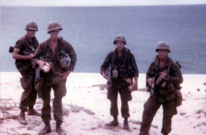 Four U.S. soldiers in full gear, standing on the beach.