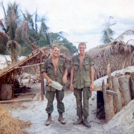 Two young U.S. soldiers standing near some huts on what may be a beach.
