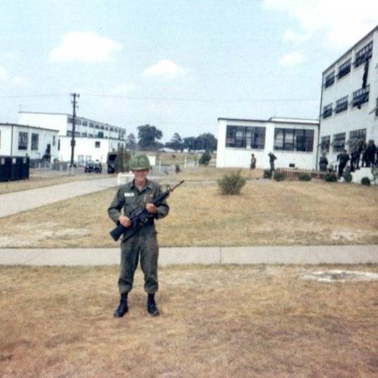 A young U.S. soldier in full gear, holding his rifle, outside some white buildings.