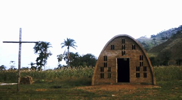 A small hut at the foot of some hills, a cross is erected in the foreground.