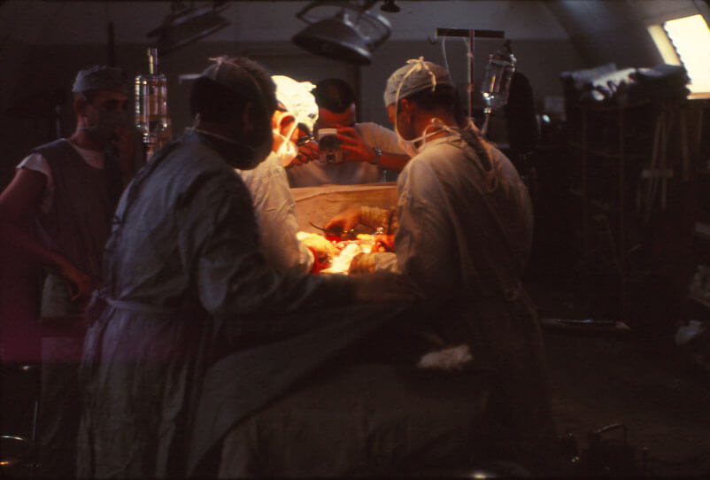 Operating Room consisting of four men standing around a table operating on a wounded soldier, another person stands at the head of the table with a camera photographing the operation.