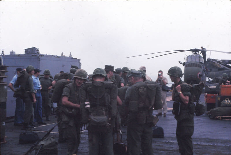 A group of Marines with packs standing on the deck of a ship with a helicopter in the background.