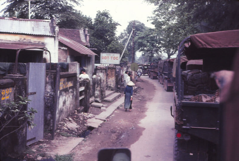 Convoy trucks passing through a village, a few Vietnamese people standing off to the side.