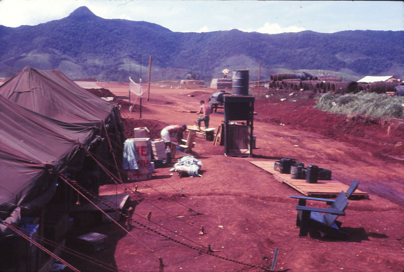 A tent to the left, a wooden stand and barrel - presumably the shower - at center, mountains in the background.