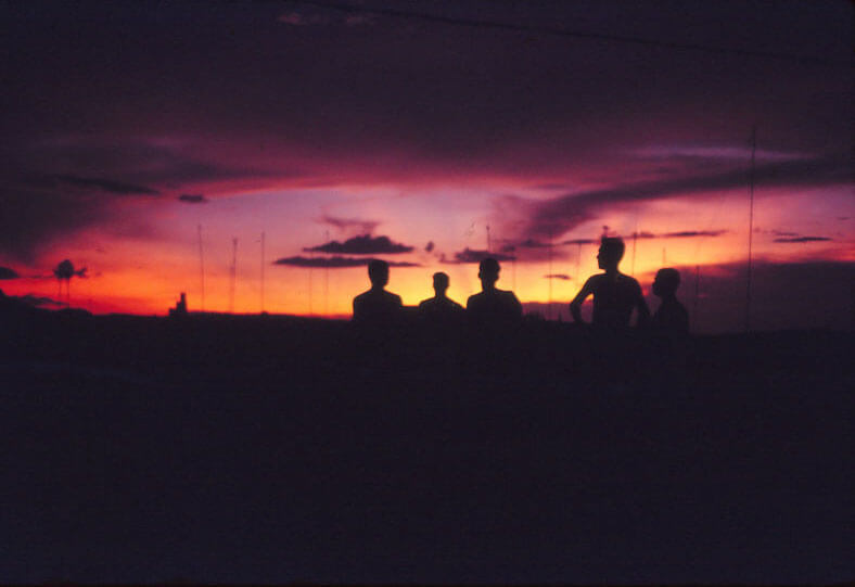 Silhouettes of three men and radio antennas against a pink, yellow, and orange sky.