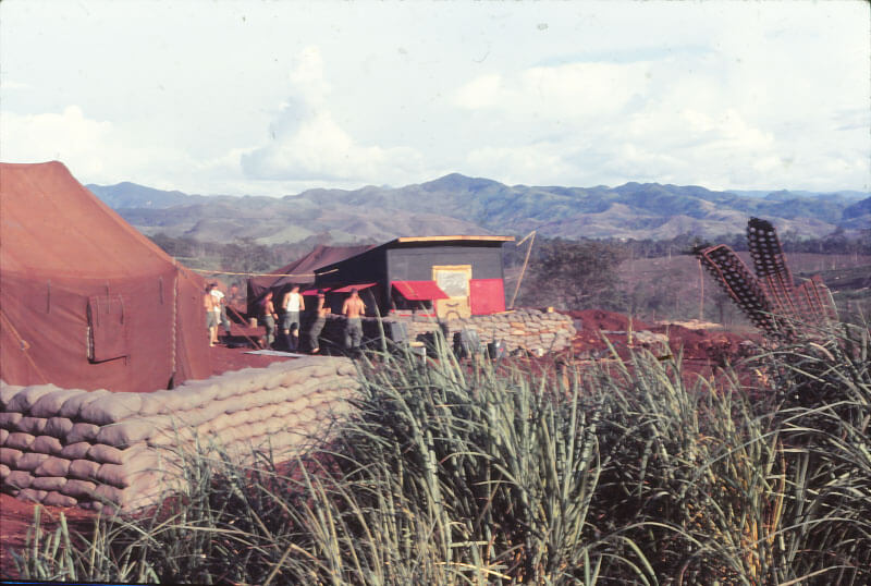 A tent surrounded by sandbags, a group of soldiers stand near a metal building, tall grass in the foreground and mountains in the background.