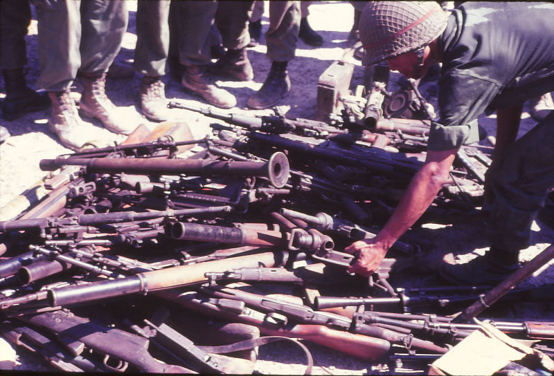 A pile of semi-automatic, automatic, rocket launchers and other assorted weapons, one soldier reaching down toward the weapons.