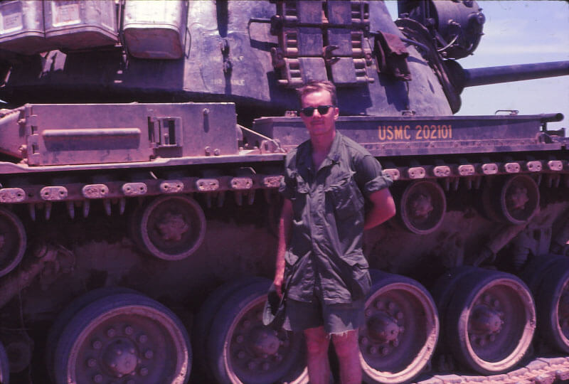 Soldier in fatigues and shorts wearing sunglasses standing in front of a tank.
