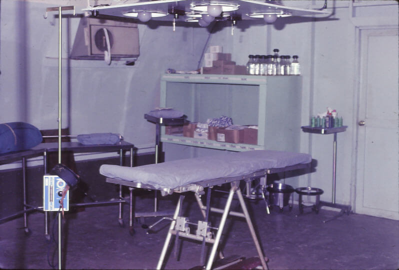 A surgical suite with a mobile gurney, lights set up over it and storage units in the background.