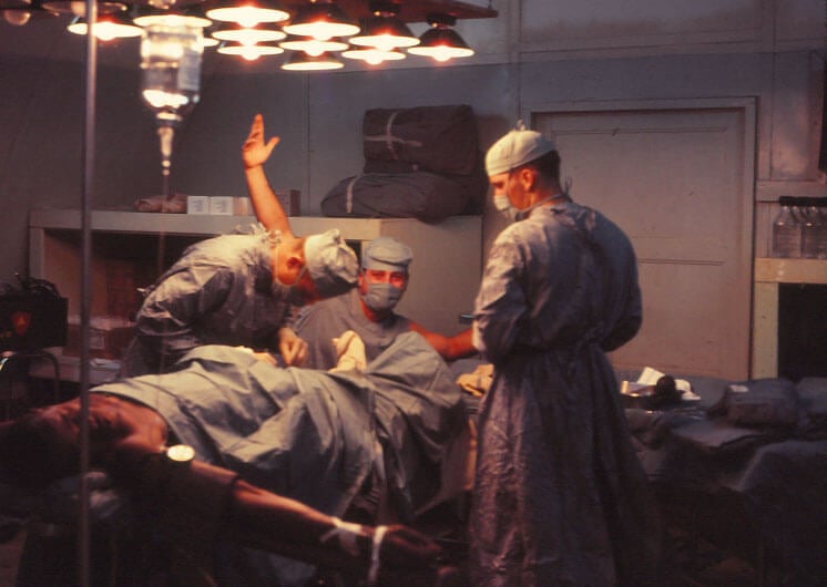 Three men in scrubs performing surgery on a wounded soldier. One man looks at the camera and holds his arm up.