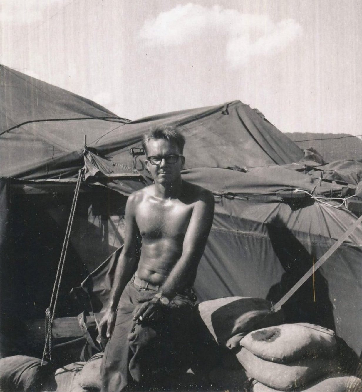 Young, shirtless man sitting on a sandbag wall holding a cigarette, behind him is a canvas tent.