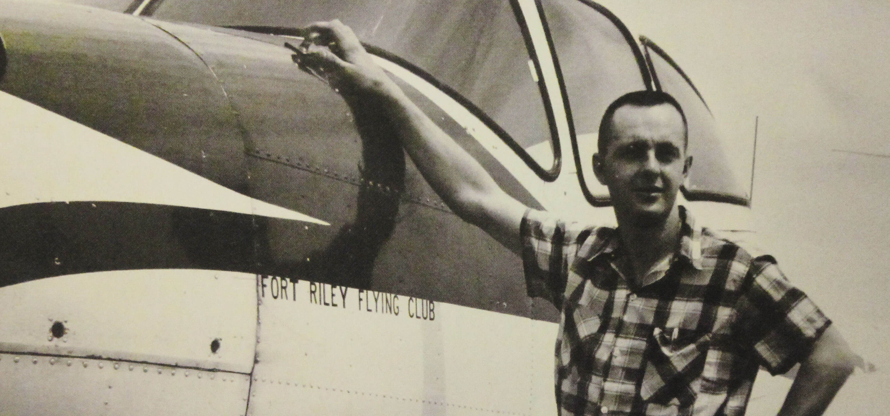 A young man in civilian clothes leaning up against a plane with the text "Fort Riley Flying Club" on its side.