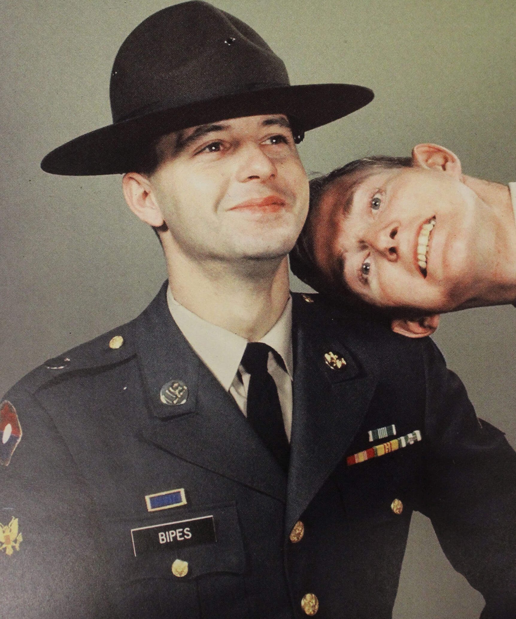 A portrait of a young soldier trying to keep a straight face while a second man leans into the frame with a grin.