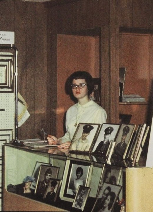 A young woman behind the desk of a photo shop, making notes on a clipboard.