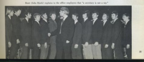 A row of young men in suits, looking to the center figure. Caption reads: "Bratt (John Bjork) explains to the office employees that "a secretary is not a toy."