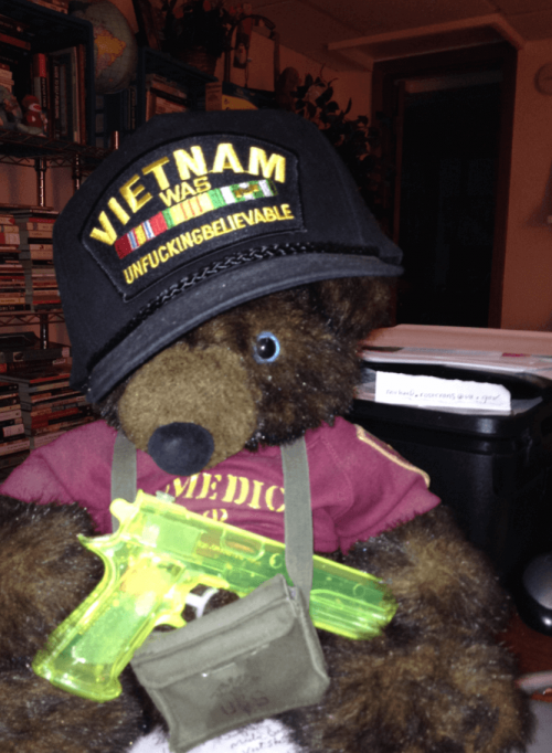 Teddy bear wearing a medic shirt, a ball cap that reads "Vietnam was unfuckingbelievable" and holding a green water pistol.